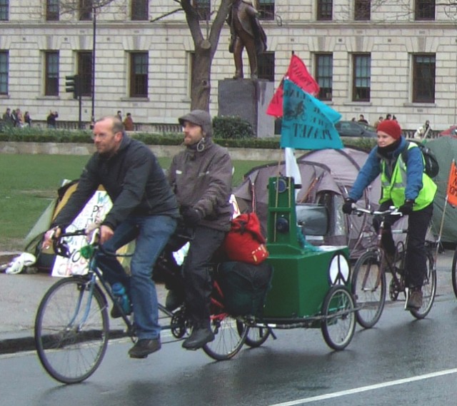 Despite rain, snow, low temperatures and wind 17 intrepid cyclists set off from London to the Atomic Weapons Research Estabishment at Aldermaston in Berkshire. Taking three days we had lots of fun & games on the way. We left on the Saturday arriving early on the Monday for the Hands Around the Base Demonstration.
Here Tim, Dan & Alida cycling past Brian Haw's encampment on Parliament Square.